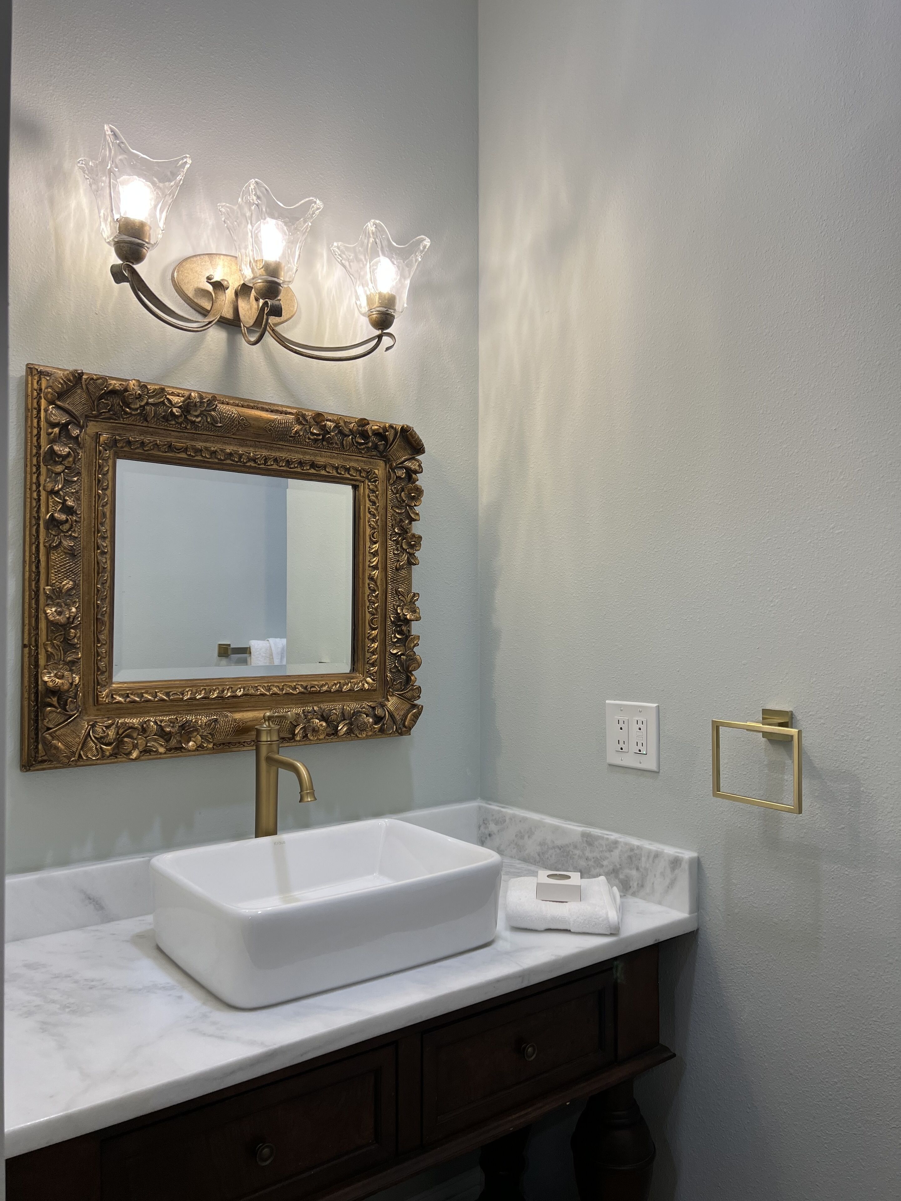 A bathroom with a white sink, a gold-framed mirror, and a wall-mounted light fixture.