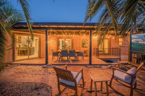 An outdoor patio area with a dining table and chairs, a kitchen visible through glass doors, and a brick wall with a painting. - Vista Velo has a Botanical Garden w Spa & Lap Pool (Tucson)