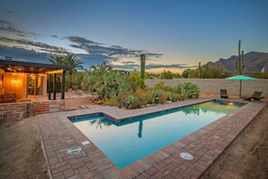 A rectangular swimming pool with a brick border, surrounded by a patio area with outdoor furniture and a covered seating area. - Vista Velo has a Botanical Garden w Spa & Lap Pool (Tucson)