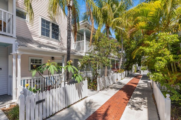 A row of white houses with a red pathway, surrounded by lush greenery and palm trees.