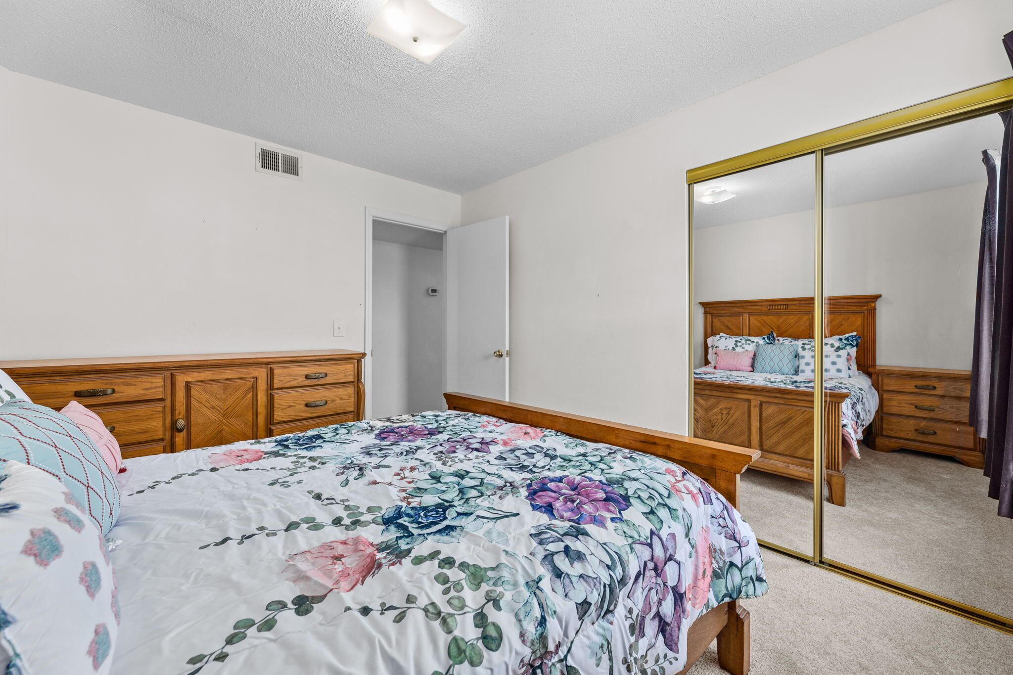 Une chambre avec un grand miroir, une tête de lit en bois, une couvre-lit à motifs floraux et une commode en bois.