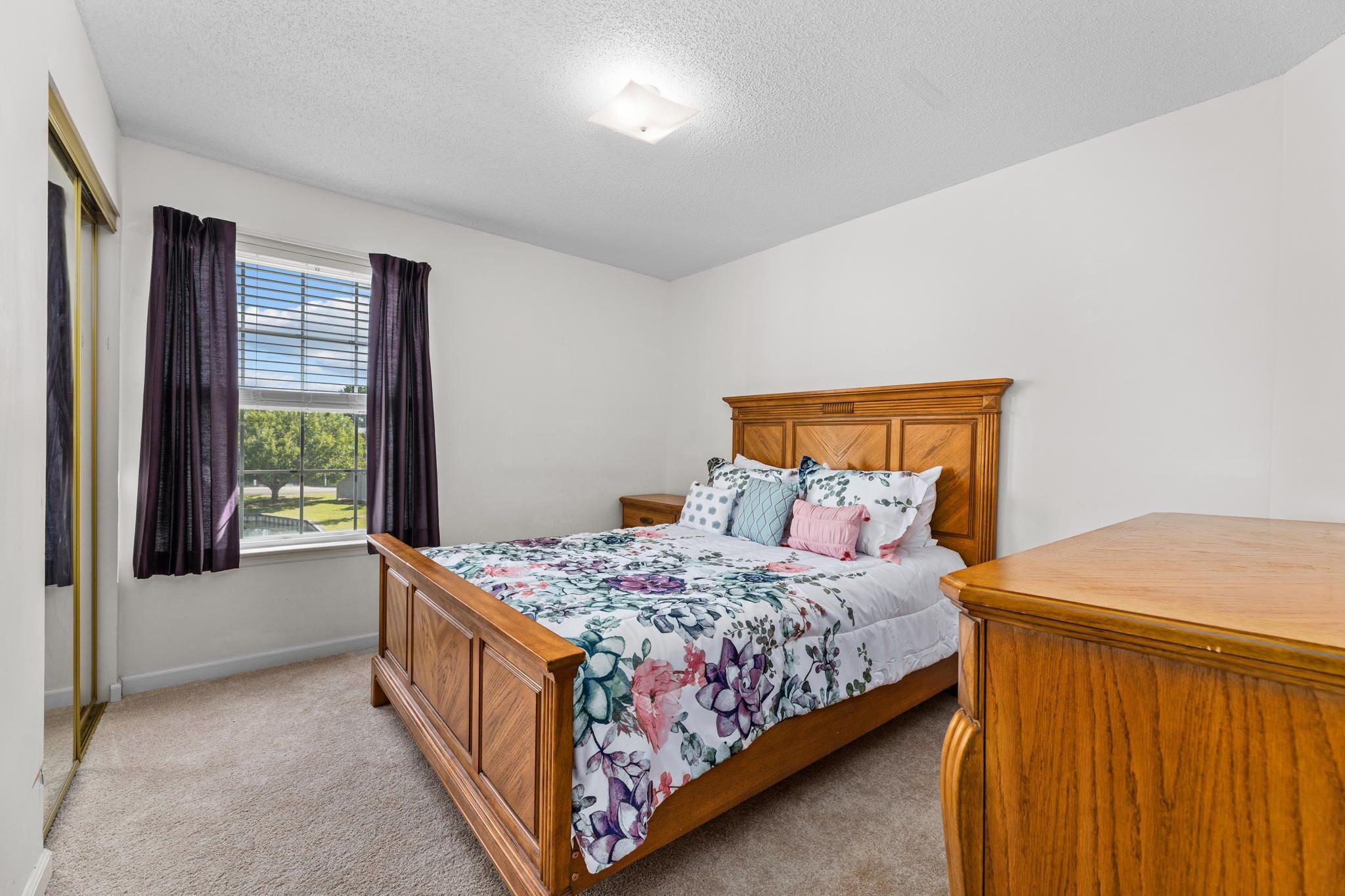 A bedroom with a wooden bed, a nightstand, and a dresser. There is a window with curtains and a view of greenery outside.
