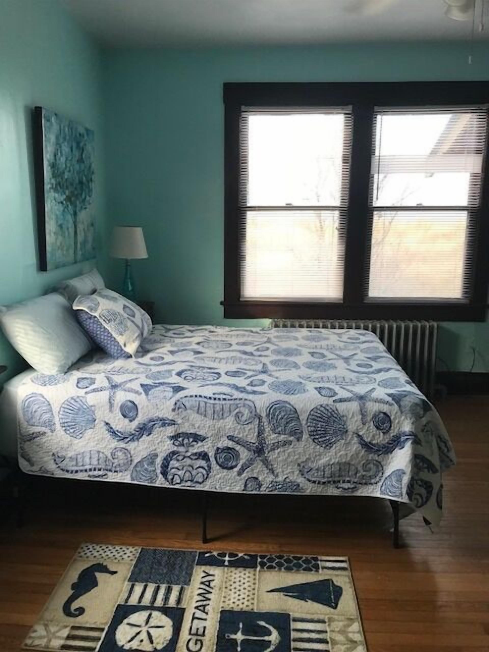 Une chambre à coucher avec un lit, une fenêtre avec des stores, une lampe et un tableau au mur.