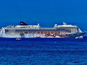 A large cruise ship with multiple decks and lifeboats visible, sailing on a deep blue ocean.