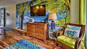 Une pièce avec un plancher en bois, une commode en bois, une chaise en osier avec un coussin vert, une fresque murale représentant des palmiers et un ventilateur de plafond.