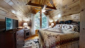 A wooden cabin bedroom with a bed, dresser, TV, and a ceiling fan.