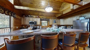 A wooden cabin interior with a stone island, wooden chairs, a refrigerator, and a window with blinds.