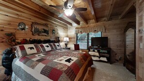 A log cabin bedroom with a bed, a ceiling fan, a television, and a window with blinds.