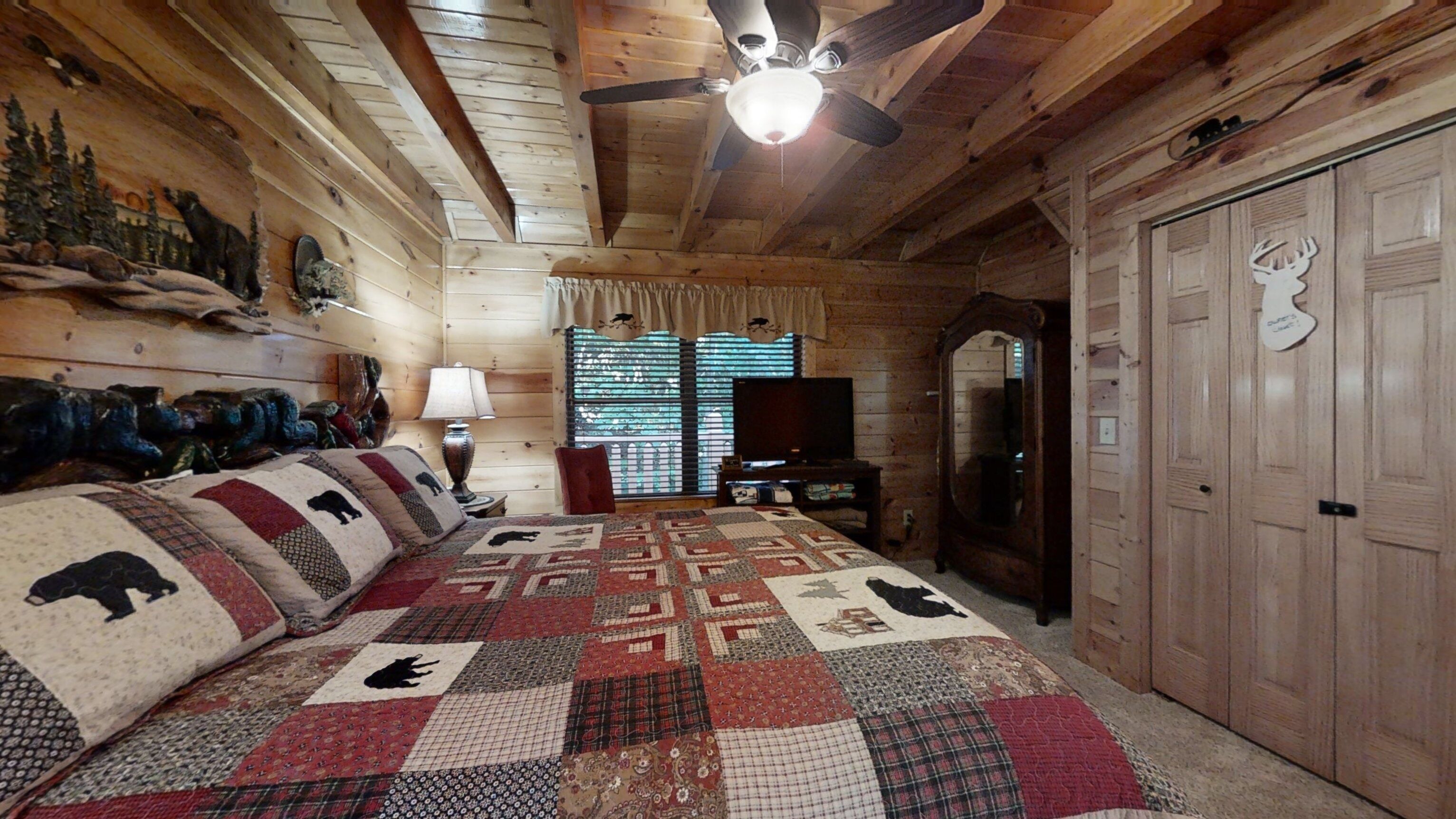 Ein Schlafzimmer in einer Blockhütte mit einer gesteppten Bettdecke mit Tiermotiven, einem Deckenventilator, einem Fernseher und einem Spiegel.