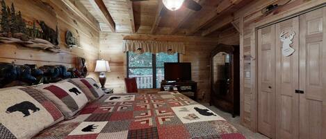 Ein Schlafzimmer in einer Blockhütte mit einer gesteppten Bettdecke mit Tiermotiven, einem Deckenventilator, einem Fernseher und einem Spiegel.