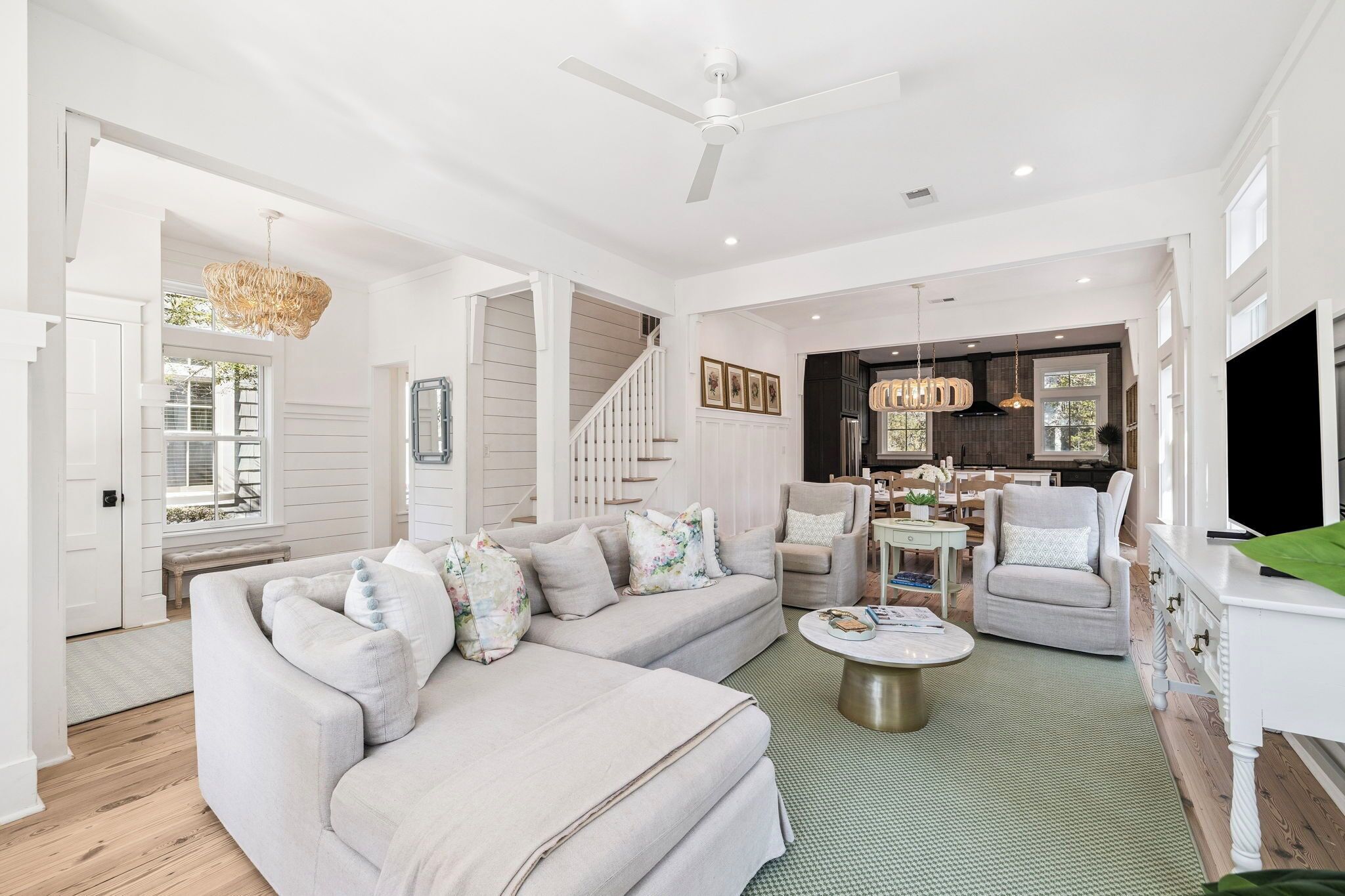 A spacious living room with a large sectional sofa, a coffee table, and a TV. There is a staircase leading to another level, and a dining area with chairs and a table.