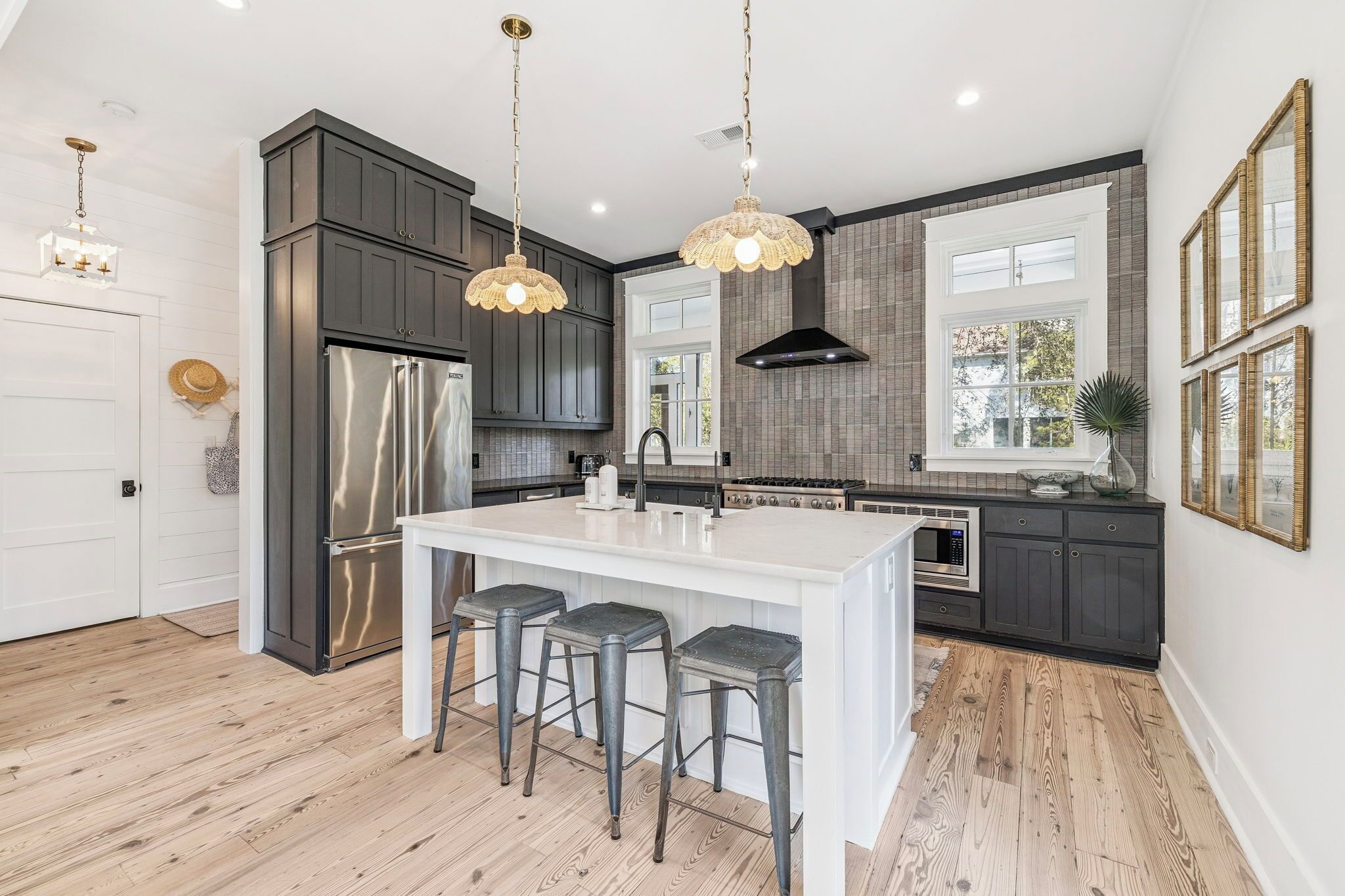 A modern kitchen with dark cabinets, a stainless steel refrigerator, a white island with bar stools, pendant lighting, and a large window.