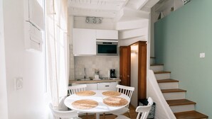 Une cuisine moderne avec des meubles blancs, un four à micro-ondes et une machine à café. On y trouve une table à manger avec quatre chaises et des sets de table en fibres tressées. Un escalier en bois mène à un palier.
