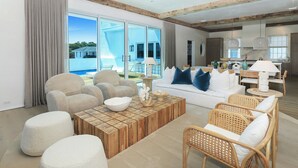 Una sala de estar moderna con un ventanal grande, un sofá, sillones, una mesa de centro y una cocina visible a través de puertas de cristal.