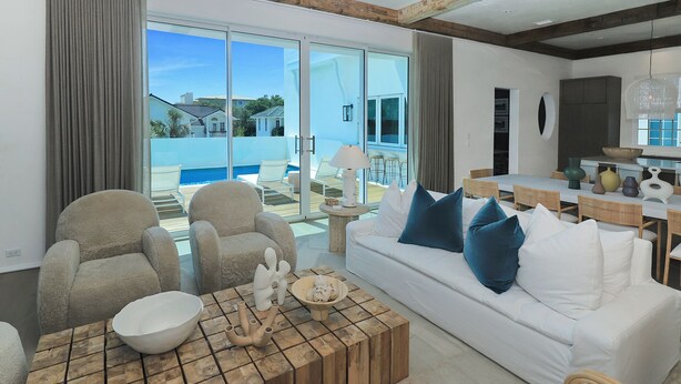 Amplia sala de estar con un sofá blanco, sillones color beige y una mesa de centro de madera. Se observa un comedor con mesa blanca y sillas de mimbre al fondo.