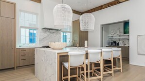Una cocina moderna con una isla de mármol, taburetes de madera, luces colgantes y vistas al exterior a través de amplias ventanas.
