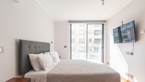 Un dormitorio moderno con una cama grande, un televisor de pantalla plana en la pared y un ventanal corredero que da a un balcón exterior.