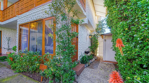Un bâtiment à l’extérieur texturé, doté d’un balcon en bois et d’un jardin où poussent différentes plantes, ainsi qu’une fleur ornementale.