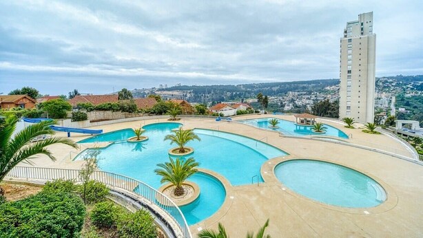A large outdoor swimming pool with multiple sections, surrounded by greenery and a tall building in the background. - Blue Oasis 2BR W/ Pool, Parking & Gym (Viña del Mar)