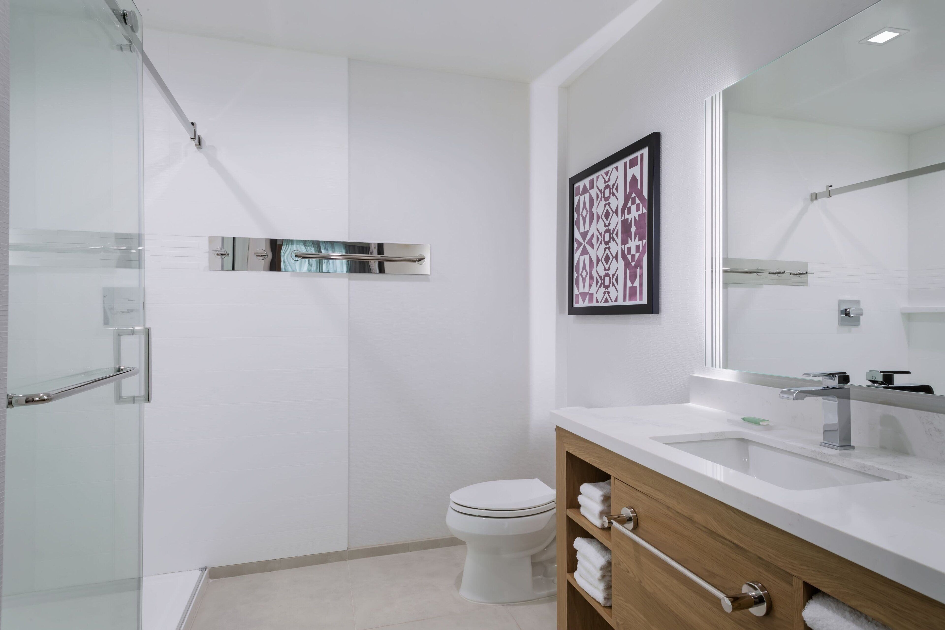 Une salle de bain moderne avec une douche à parois de verre, des toilettes blanches, un meuble-lavabo en bois avec un grand miroir et une œuvre d’art encadrée au mur.