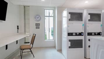 A laundry room with four stacked washing machines, a chair, a clock, and a window with blinds.