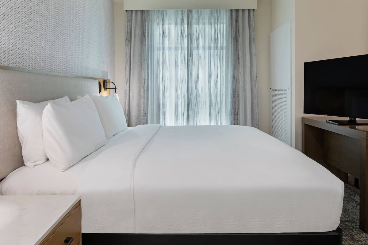 A neatly made bed with white linens, a headboard with a light fixture, a television on a wooden stand, and a window with sheer curtains.