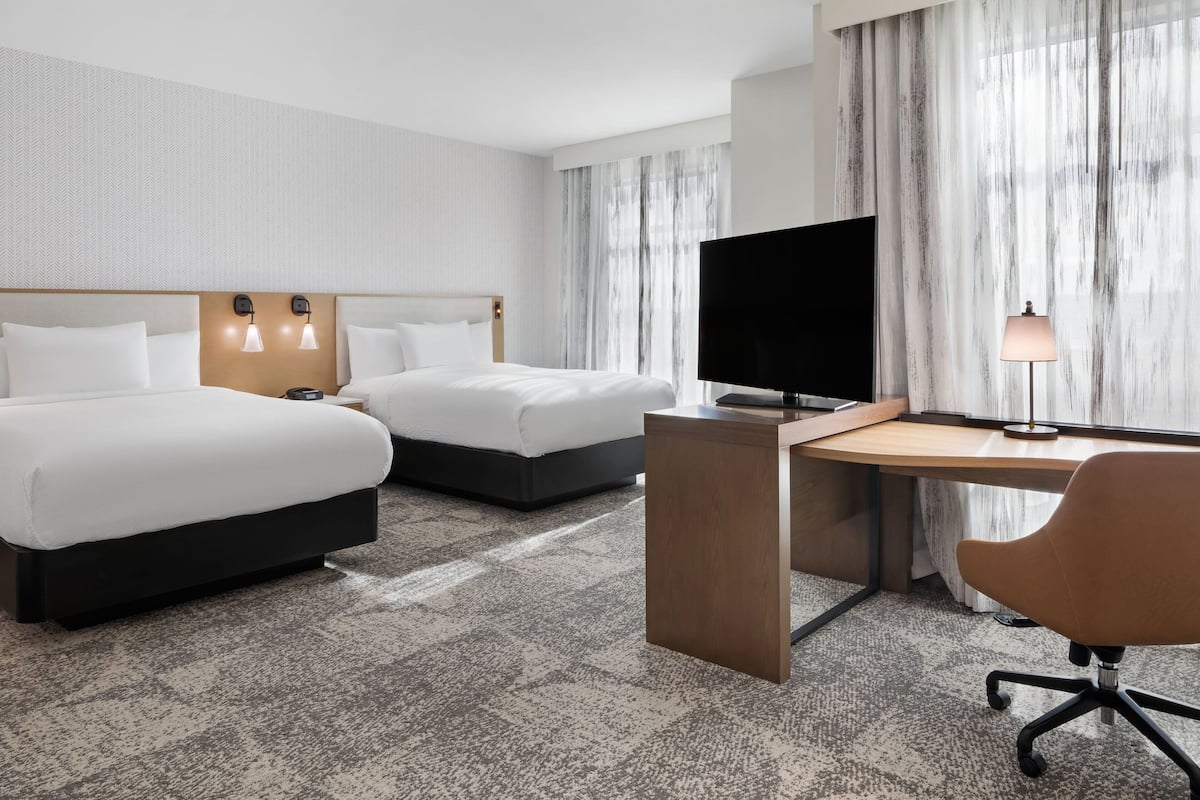 A hotel room with two beds, a desk, a TV, and a lamp.
