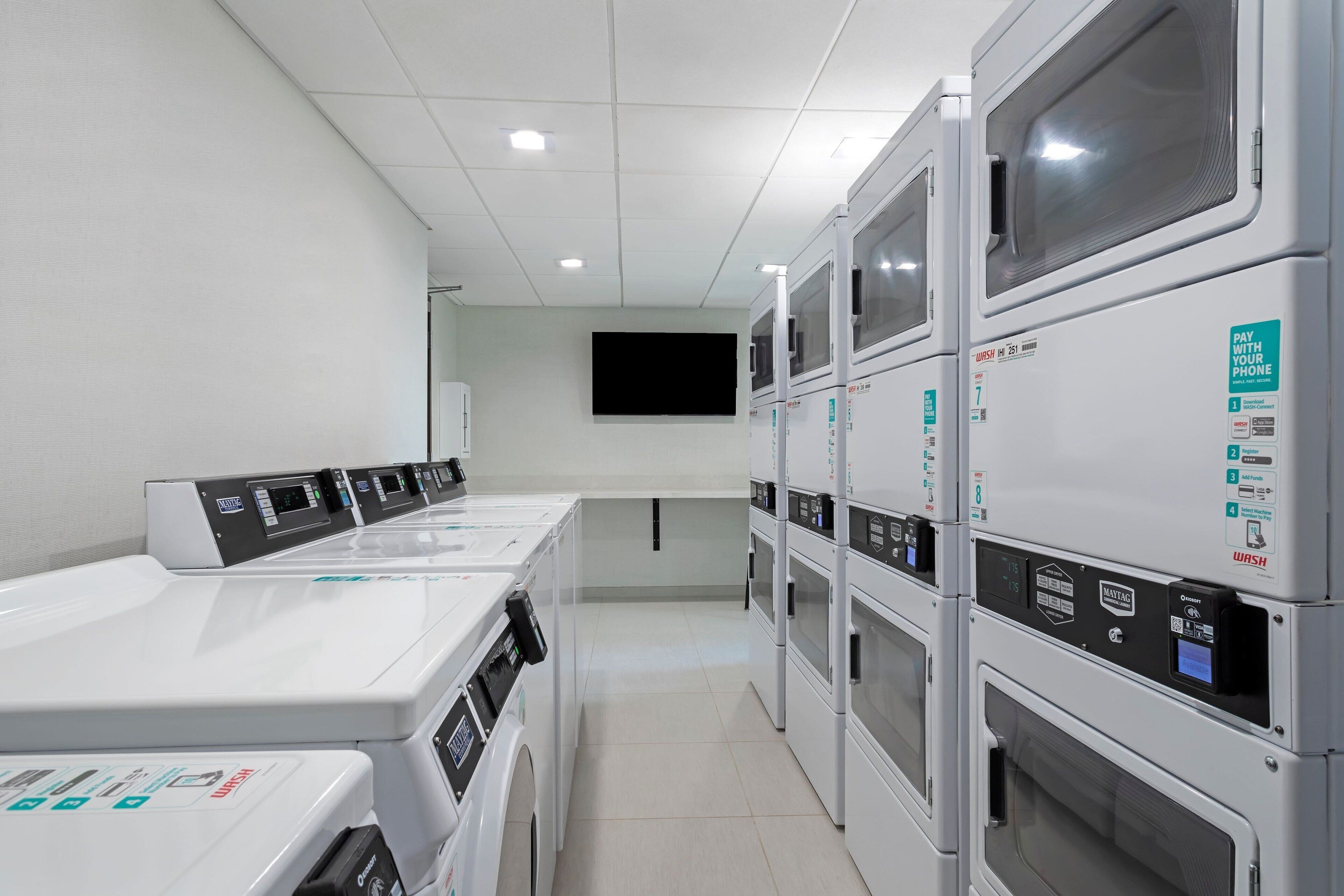 Une buanderie équipée de plusieurs machines à laver et de plusieurs sèche-linge, d’un téléviseur à écran plat fixé au mur et d’un panneau indiquant que le paiement peut se faire par téléphone.