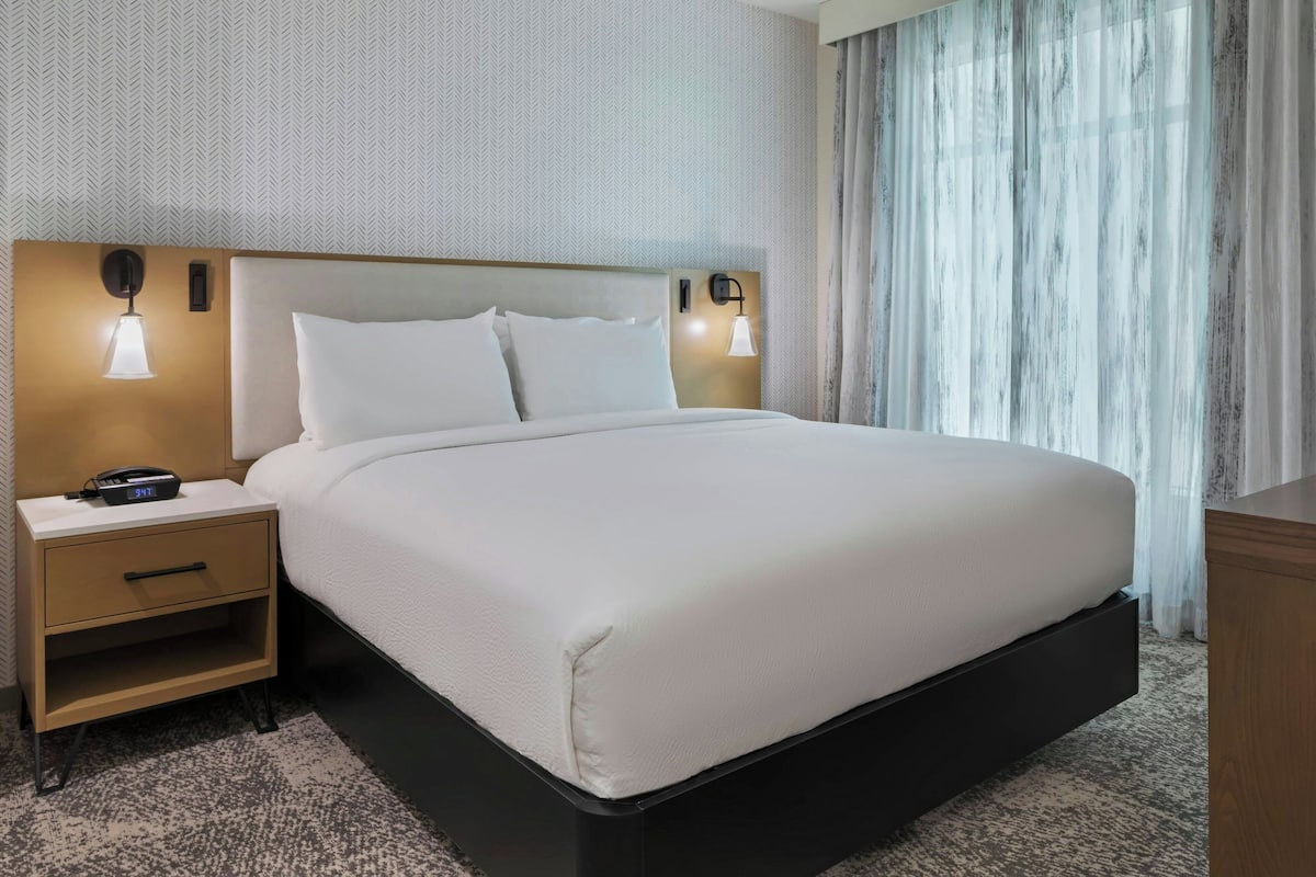 A neatly made bed with white linens, a wooden nightstand with a lamp, and a small drawer.