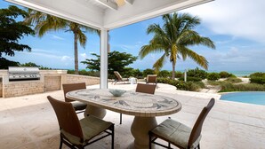 An outdoor patio with a round table and chairs, a grill, and a view of the ocean and palm trees. - LUXURY BEACHFRONT Villa on EXQUISITE BEACH, crystal WATER. IN SANDCASTLE ESTATES (Sandy Point)