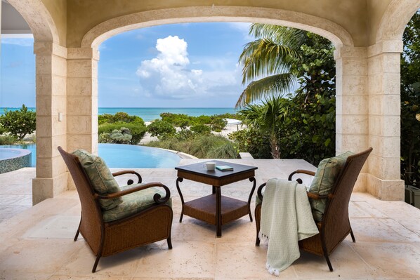 Un espacio de estar al aire libre cubierto, con sillas de mimbre y una mesa, con vistas a una piscina y una playa.