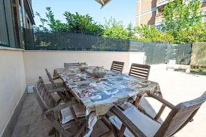 Una veranda esterna con un lungo tavolo di legno, sedie e una tovaglia fantasia.