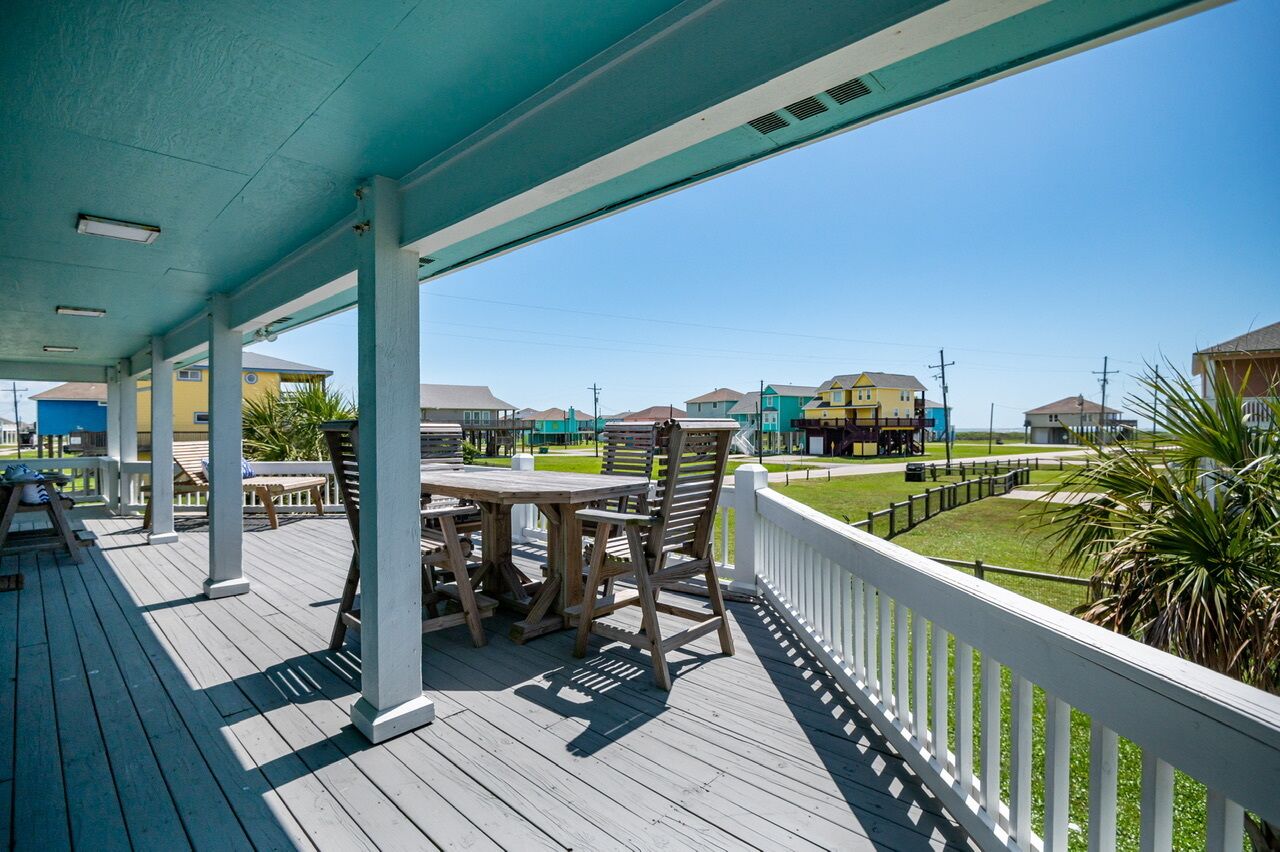 A covered outdoor area with a wooden table and chairs, a white railing, and a view of colorful buildings in the background.