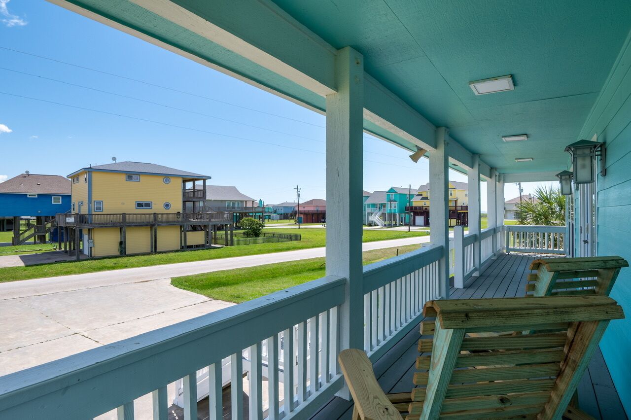A covered porch with wooden benches, a view of a street with houses, and a clear sky.