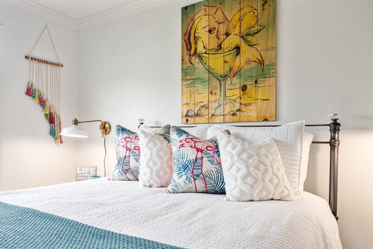 Un lit bien fait, recouvert d’une couette blanche et bleue, une lampe fixée au mur et, au-dessus, un tableau abstrait aux couleurs vives.