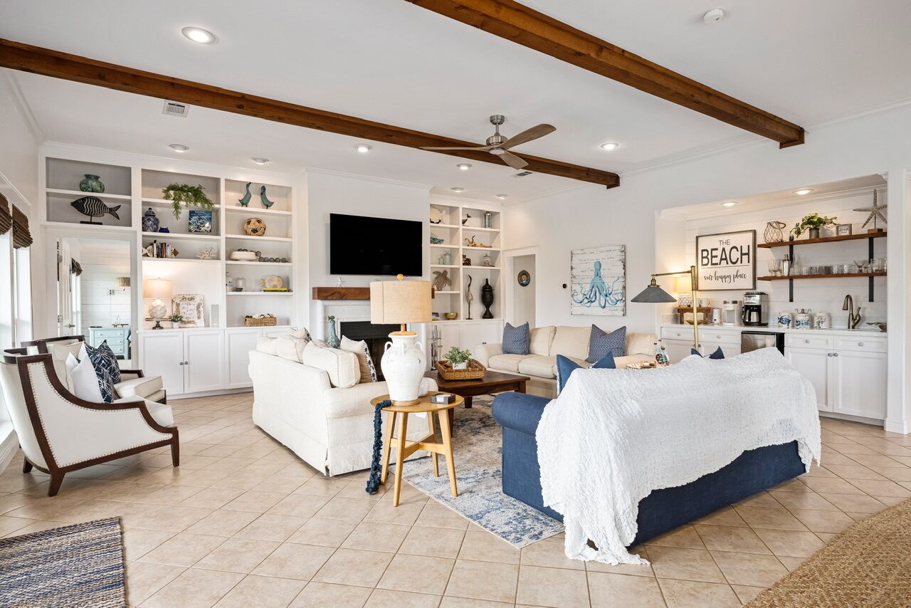 A spacious living room with a white sofa, blue armchair, and a coffee table. There are shelves with decorative items and a television mounted on the wall.