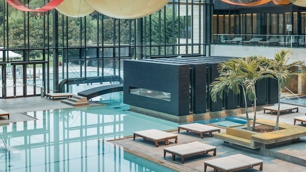 Un espace piscine intérieur moderne, doté de parois en verre transparent, de chaises longues et d’un élément décoratif central avec de l’eau.