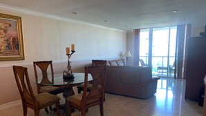 A dining area with a round glass table and wooden chairs, a sofa, and a painting on the wall. - Entire condominium with a pool view, steps from the beach (Sunny Isles Beach)