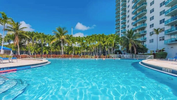 A large, clear swimming pool surrounded by palm trees and a tall building with balconies. - Entire condominium with a pool view, steps from the beach (Sunny Isles Beach)
