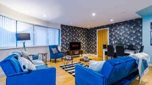 Un salon moderne avec des canapés bleus, un téléviseur et un coin repas agrémenté d’un mur à motifs.