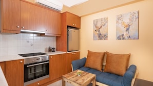 A compact kitchen with wooden cabinets, a stove, and a refrigerator. There is a blue sofa with two pillows and a small wooden table with a plant. - Athens Riviera Golf Apartment (Γλυφάδα)