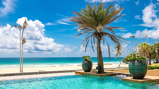 Uma piscina com águas cristalinas azuis, palmeiras e uma praia ao fundo.