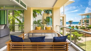 Uma varanda com mesa e banco de madeira, rodeada por plantas tropicais e vista para o oceano.
