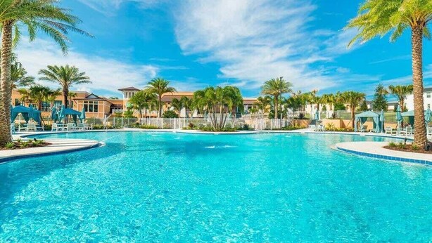 Una gran piscina al aire libre rodeada de palmeras y sillas de descanso, con edificios al fondo.