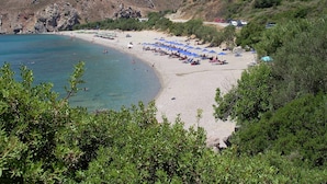 A beach with a row of blue umbrellas, rocky hills in the background, and a few buildings on the hillside. - Metatulia 3 villa,sea views,pool,Souda,Plakias (Plakias)