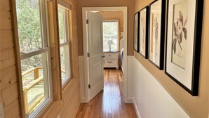 A hallway with wooden walls, a wooden floor, and a door leading to a bedroom. There are framed botanical prints on the wall. - Cozy Mountain Cabin Nestled on 3.5 Acres - Multi-Tier Deck, Creek Access! (Clyde)