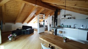 A spacious loft with a high ceiling, wooden beams, a kitchen with white cabinets, a dining area with a wooden table, and a living room with a sofa and a coffee table. - Cozy stay in the heart of Mondovi (Mondovi')