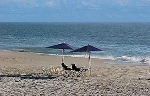 A beach with two umbrellas, a chair, and a small table.