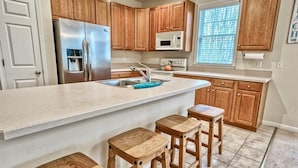 A kitchen with wooden cabinets, a stainless steel refrigerator, a microwave, and a sink. There are three wooden stools and a countertop. - Bear Trap Dunes * 4BR Bethany Beach Luxury Home (Ocean View)