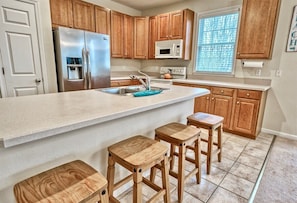 A kitchen with wooden cabinets, a stainless steel refrigerator, a microwave, and a sink. There are three wooden stools and a countertop.